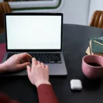 Tangan mengetik di laptop di sudut kerja rumah dengan kopi dan buku di sisi meja, menggambarkan penggunaan internet untuk bekerja dari rumah
